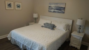 Een net opgemaakt bed met een witte deken en een grijze kussen, twee ingelijste foto's aan de muur, en een schilderij boven het bed.