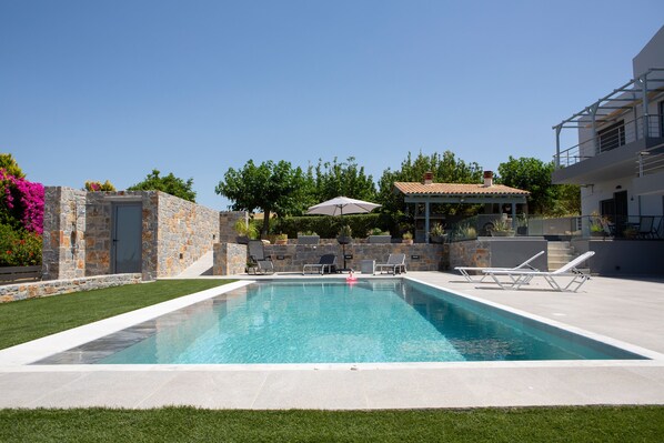 A rectangular swimming pool with a stone wall on one side, a covered seating area with umbrellas, and a white building with a balcony in the background.