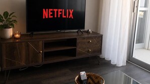 Ett modernt vardagsrum med en platt-TV som visar ”NETFLIX”, en träinredd TV-möbel, ett glasbord och en krukväxt.