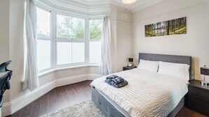 A bedroom with a large window, a bed with pillows, a nightstand, and a painting on the wall.