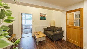 A cozy living room with a sofa, a coffee table, and a TV. There's a wooden door and a window with a view.