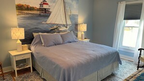 A bedroom with a large bed, two bedside tables with lamps, a nightstand, a patterned rug, and a wall mural of a lighthouse and sailboat. - 3-bedroom historic vacation home newly renovated in the middle of Oxford MD. (Oxford)