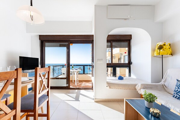A modern living room with a TV, wooden chairs, a sofa, and a balcony with a view of the sea.