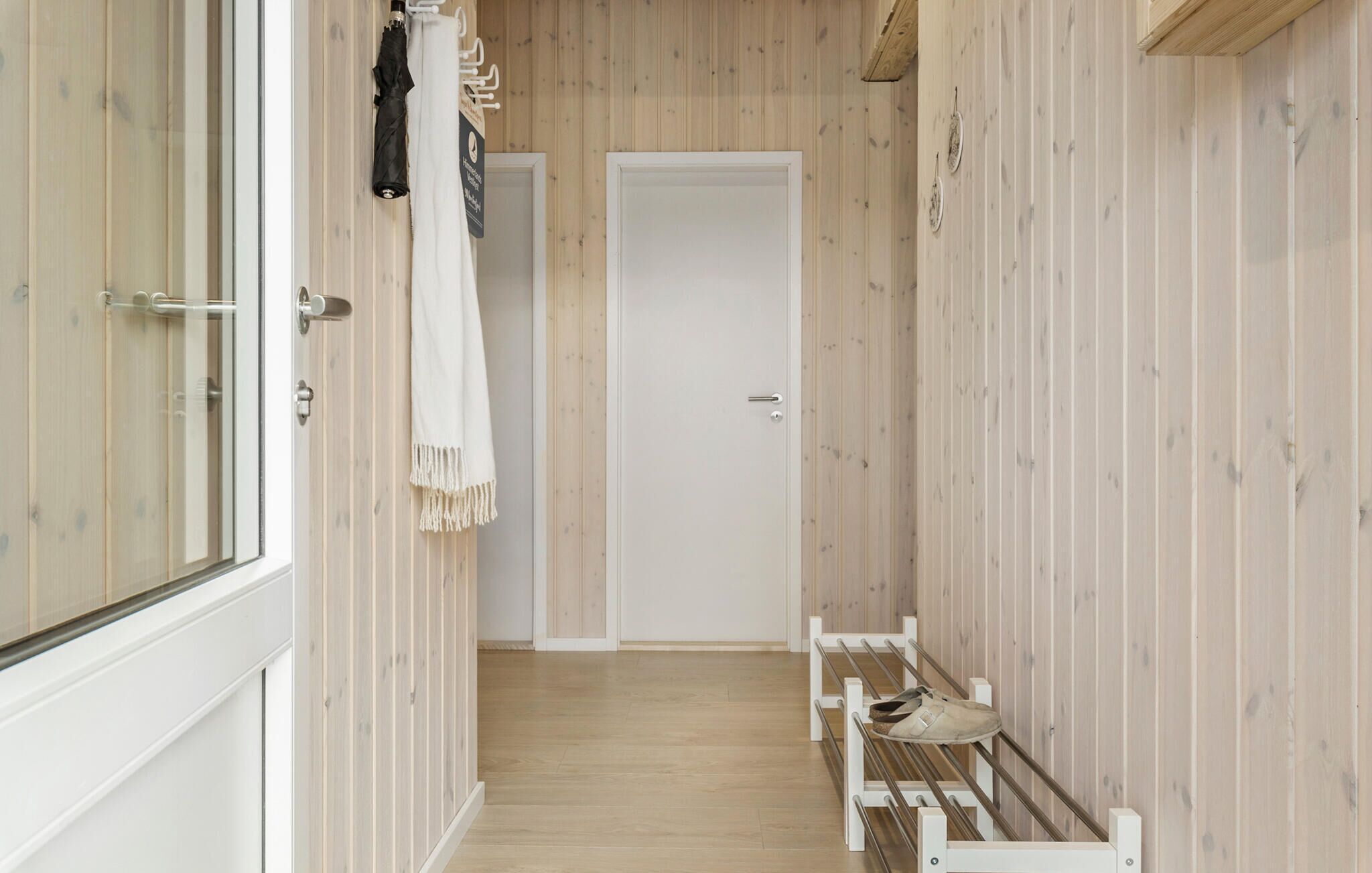A narrow hallway with wooden walls, a white door at the end, and a shoe rack with shoes.