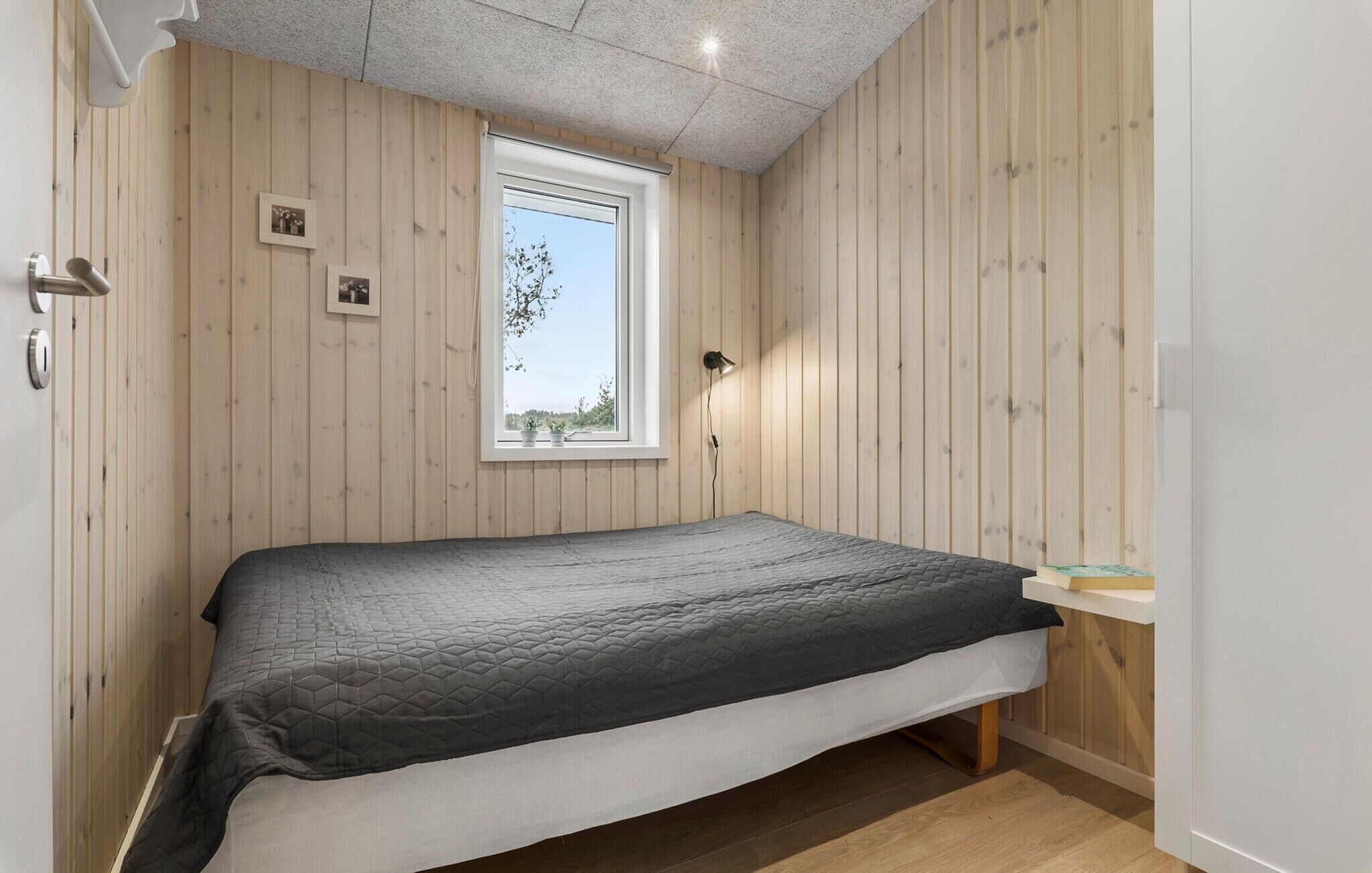 A single bed in a room with wooden paneling, a window with a view of trees, and a wall-mounted lamp.