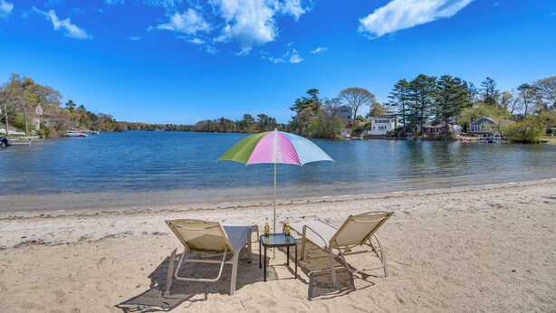 Una playa con dos sillas y una mesa bajo un paraguas colorido, con vista a un lago tranquilo y casas al fondo.