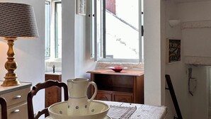Interior rústico com paredes de pedra aparente, uma mesa de jantar de madeira com um jogo americano de renda, um jarro e tigelas. Uma janela revela a vista de um prédio vermelho.