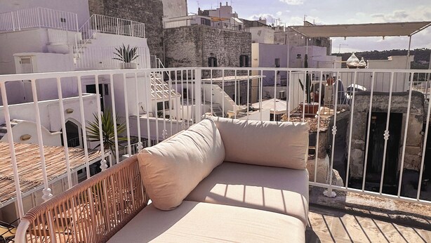 Terraço no telhado com guarda-corrimão de metal branco, sofá bege com almofadas e vista para a paisagem urbana.