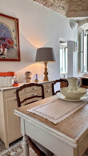 Een gezellige keuken met een houten tafel, een kantkleed en een lamp. Aan de muur hangt een schilderij en er is een raam met gordijnen.