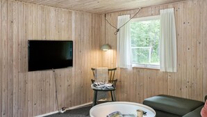 A room with wooden walls, a flat-screen TV mounted on the wall, a window with white curtains, a green chair with a striped towel, a small round table with a magazine and a cup, and a green sofa. - Lovely home in Nykøbing Sj with WiFi (Nykøbing Sj)