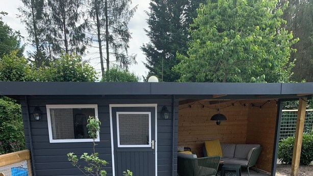 A small, dark-colored building with a door and window, a patio area with seating, and a brick pathway leading to the entrance. - Charming dog-friendly lodge in, Veluwe. Enjoy the nature by walking or cycling. (Putten)