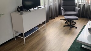 A modern living room with a flat-screen TV on a white cabinet, a grey armchair, and a green area rug. - Charming dog-friendly lodge in, Veluwe. Enjoy the nature by walking or cycling. (Putten)