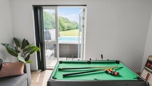 A pool table with pool cues and balls, a sofa, a potted plant, and a view of a backyard with a pool and greenery. - Awesome home in Haderslev with sauna (Haderslev)