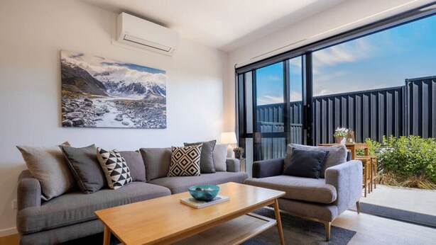 A modern living room with a grey sofa set, a wooden coffee table, and a large window offering a view of a balcony with a wooden fence and greenery. - Explore Wanaka - A Stylish Unit That Guests Adore! (Albert Town)