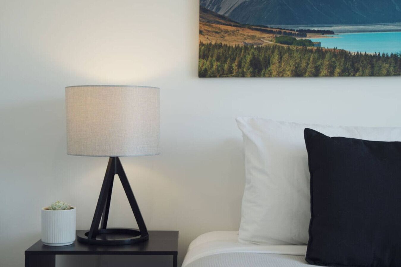 A neatly arranged hotel room with a bed, a bedside lamp, a small potted plant, and a wall-mounted painting of a mountainous landscape.