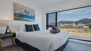 A modern hotel room with a large bed, a nightstand, a lamp, and a painting of a mountain landscape. - Explore Wanaka - A Stylish Unit That Guests Adore! (Albert Town)