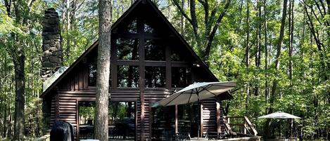 Un chalet rustique doté d’une cheminée en pierre, de grandes fenêtres et d’une terrasse en bois avec des meubles d’extérieur.