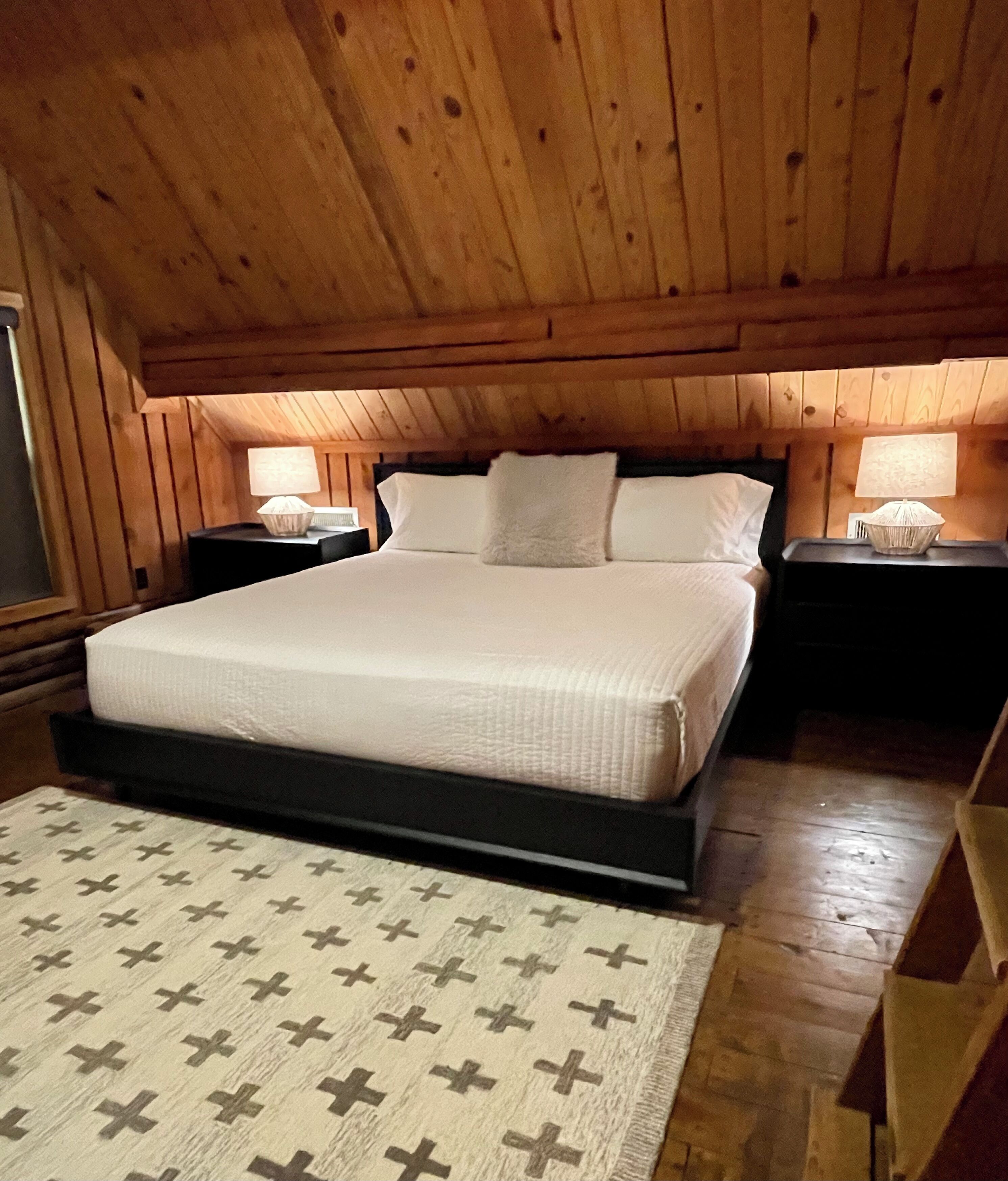 Un dormitorio con paneles de madera, una cama grande, dos mesitas de noche con lámparas y una alfombra con estampado en forma de cruz.