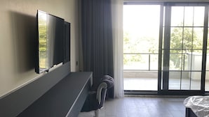A modern hotel room with a flat-screen TV mounted on the wall, a dark-colored desk, a cushioned chair, a bed with a mattress cover, and a large sliding glass door leading to an outdoor balcony. - The Reference Club Restaurant & Accommodation (Luleburgaz)