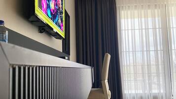 A modern hotel room with a flat-screen TV mounted on the wall, a radiator below, a chair, and a large window with sheer curtains.