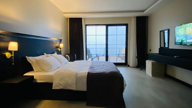 A hotel room with a large bed, a TV, a desk, and a view of the sea through a large window. - The Reference Club Restaurant & Accommodation (Luleburgaz)