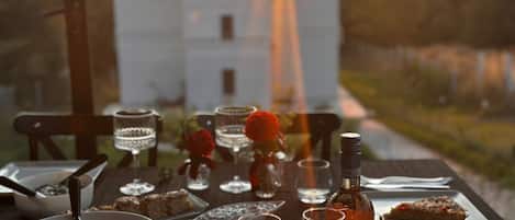 Una mesa dispuesta para una comida con copas de vino, una botella de vino Cielo y diversos platos, con vista a un atardecer y un edificio.