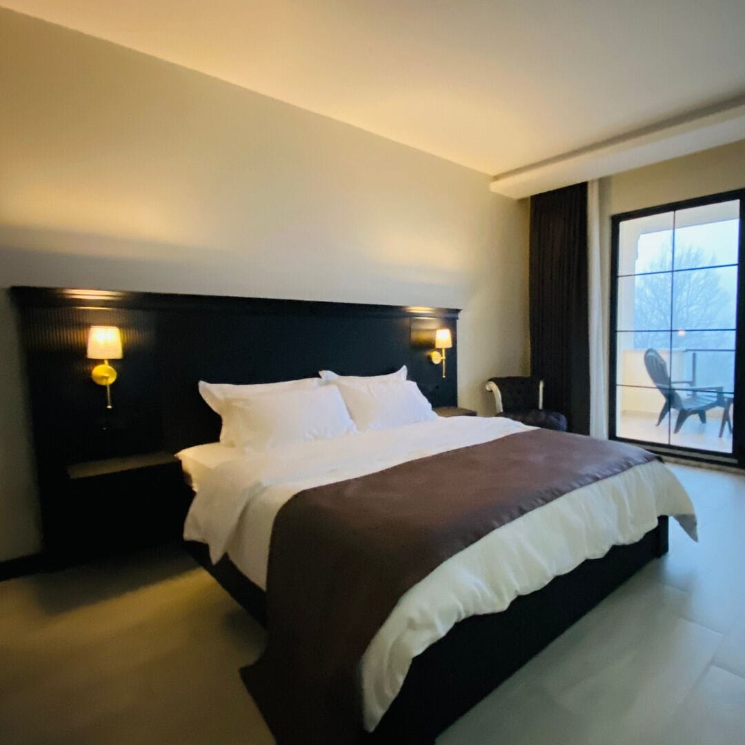 Una habitación de hotel con una cama grande, dos lámparas de noche, una silla y vistas amplias por un gran ventanal.