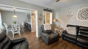 Una sala de estar con un sofá de cuero negro, un sillón reclinable de cuero y una zona de comedor con una mesa y sillas blancas. Se ve un cartel con la palabra 'welcome' en la pared.