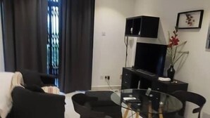 A modern living room with a sofa, a glass dining table with chairs, a TV, and a decorative plant. - Embassy garden ebs legend luxury studio  (Accra)