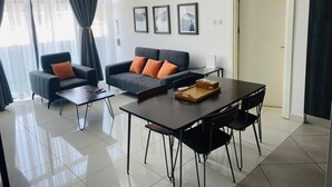 A modern living room with a sofa, two armchairs, a coffee table, and a dining table with chairs. There are three framed pictures on the wall, a door, and an air conditioning unit. - Embassy garden ebs legend luxury studio  (Accra)