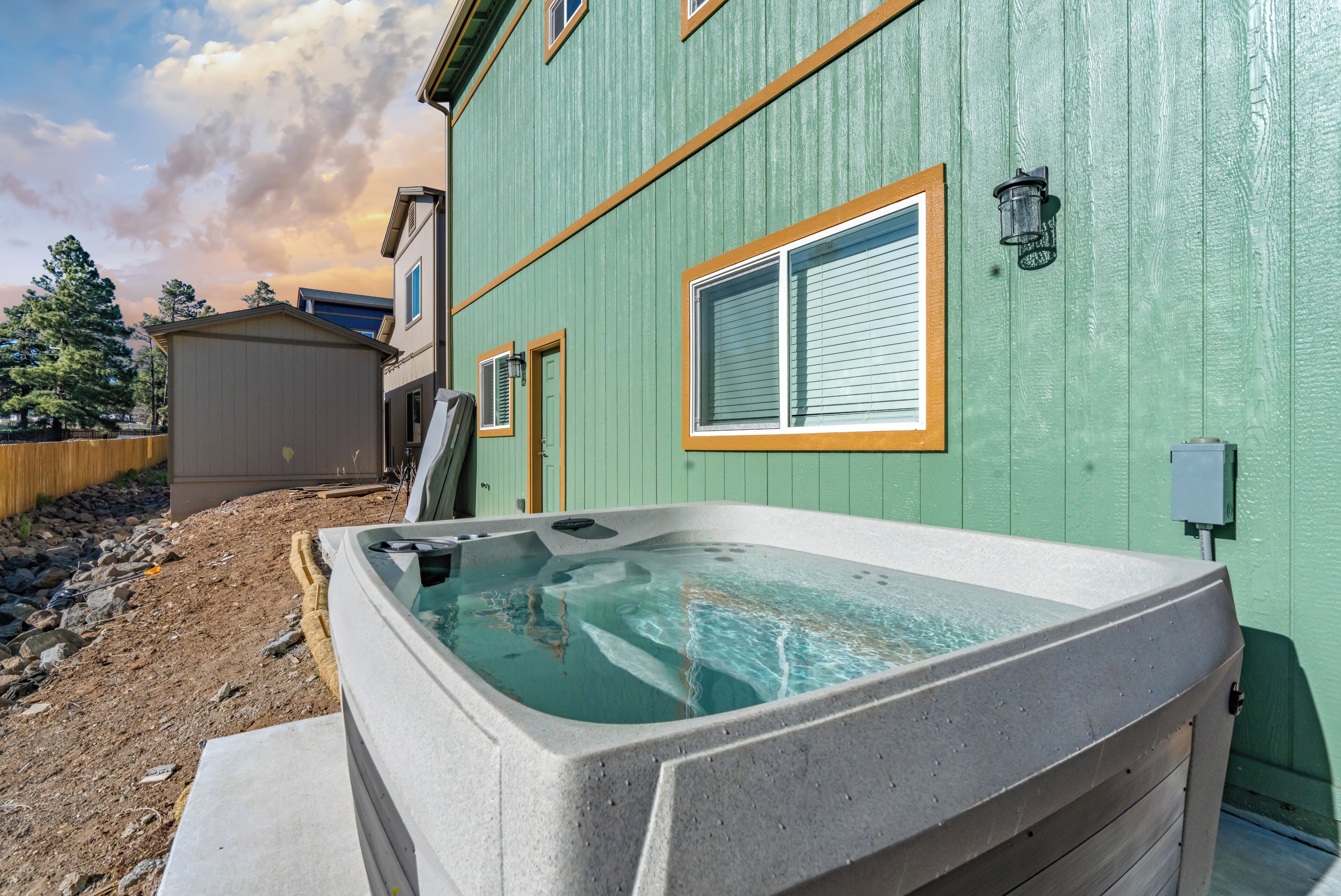 Un espace de détente avec un jacuzzi, situé devant un bâtiment verdoyant doté de fenêtres et surmonté d’une clôture en bois en arrière-plan.