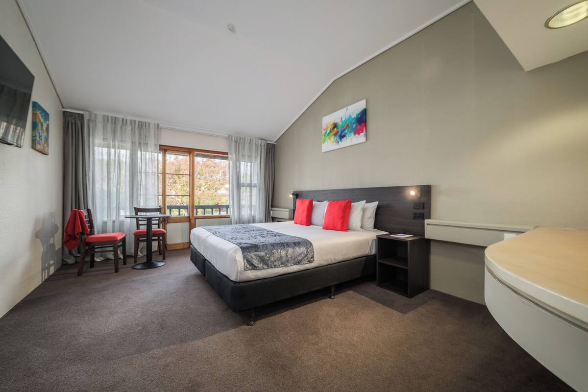 Quarto com cama, mesa de cabeceira, cadeira e janela com cortinas.