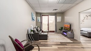 Una sala de estar moderna con un sofá gris, una mesita con una planta en maceta, un televisor de pantalla plana y un ventanal con cortinas.