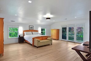 Quarto com cama, mesa de cabeceira, cômoda, sofá e escrivaninha com cadeira. Há duas janelas e uma porta que dá para uma varanda.