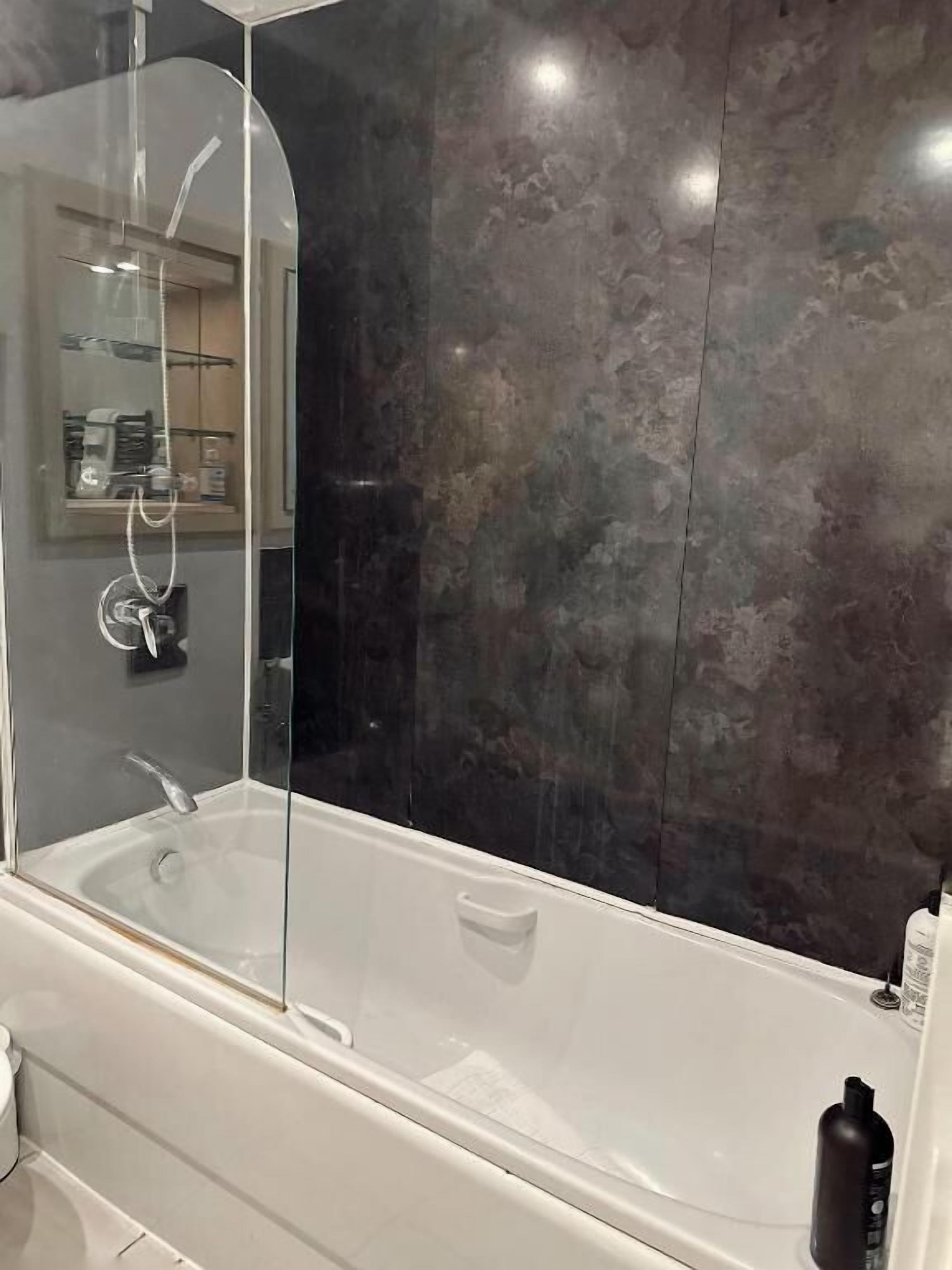 A modern bathroom with a glass-enclosed bathtub, a large mirror, and a dark textured wall.
