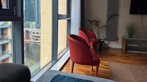 A modern living room with large windows, a view of a river, red chairs, a TV, and a plant. - Canary Wharf Apartments (London)