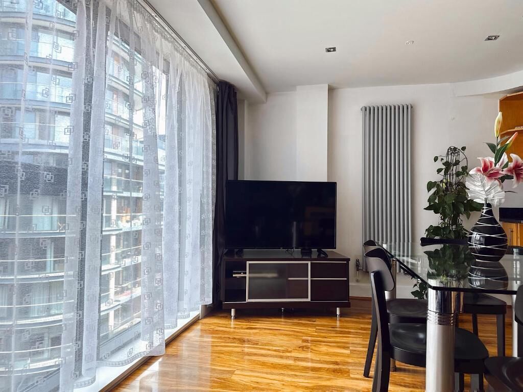 Una sala de estar moderna con un televisor de pantalla plana grande, piso de madera, una mesa de comedor de vidrio y vistas a un paisaje urbano a través de cortinas translúcidas.