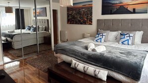 A modern hotel room with a large bed, a mirror, cityscape paintings, and a view of the city. - Canary Wharf Apartments (London)