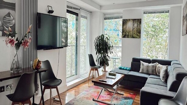 A modern living room with a sofa, chairs, a coffee table, and a TV. There are large windows and a plant. - Canary Wharf Apartments (London)