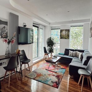 Una sala de estar moderna con un sofá, sillones, una mesa de centro y un televisor. Hay ventanas amplias y una planta.