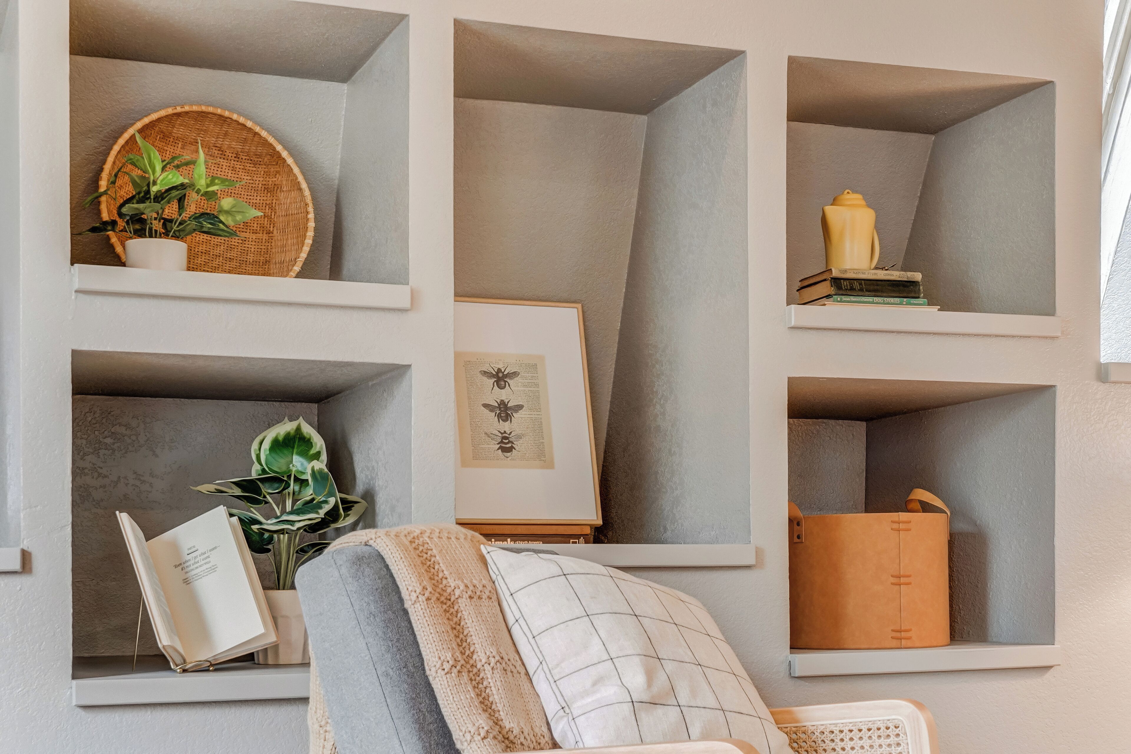 Una habitación con varios estantes incorporados que contienen objetos diversos como libros, un cuadro enmarcado, una planta en maceta y una cesta de mimbre.