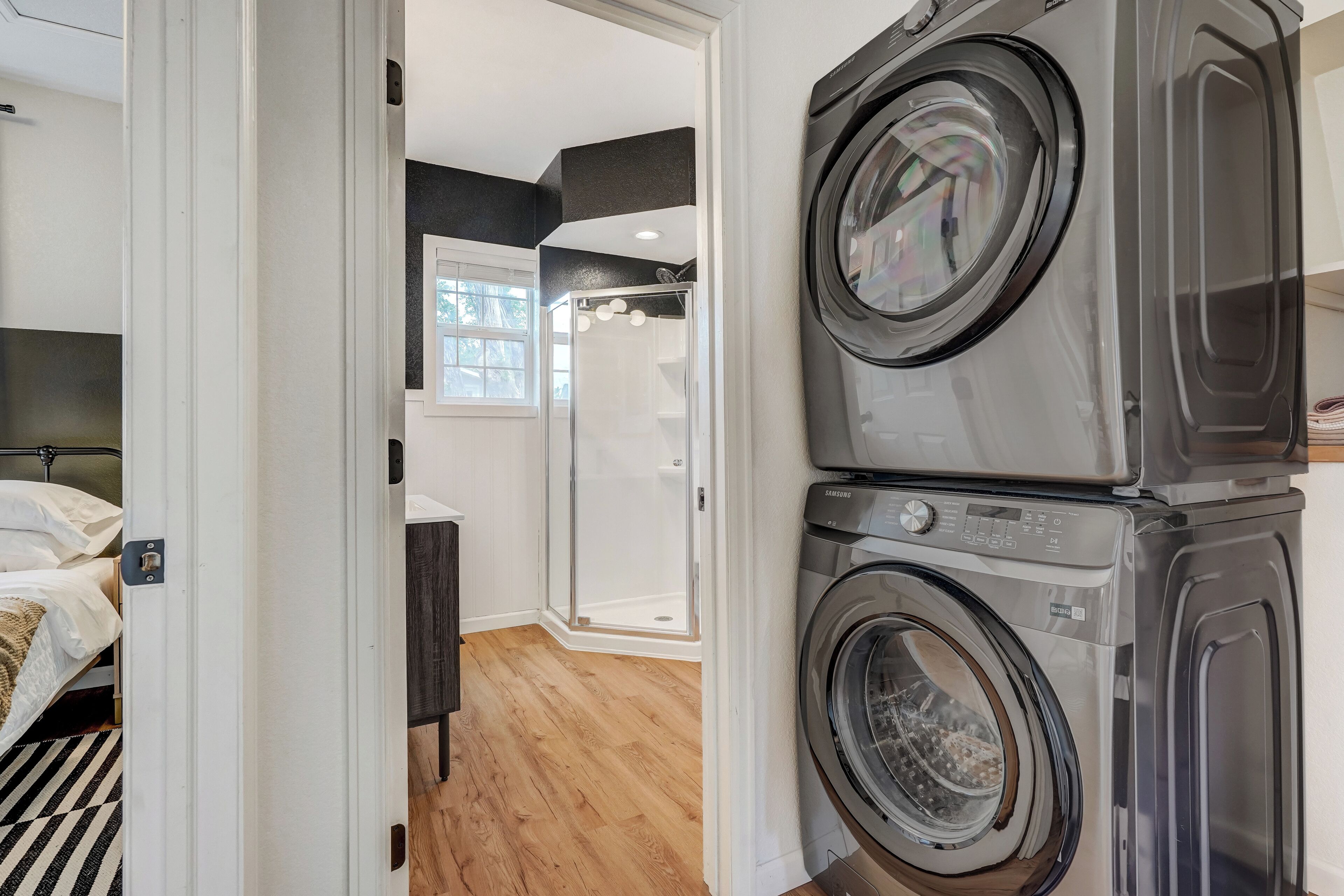 A laundry area with stacked washers and dryers, a shower with glass doors, and a bed with a striped rug.