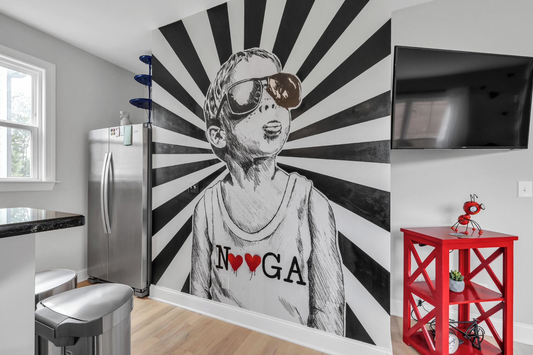 Una cocina moderna con un refrigerador de acero inoxidable, una mesita auxiliar roja con un reloj y un mural en la pared.