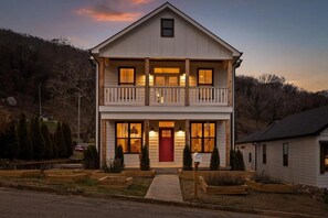 Une maison à deux étages avec un porche à l’avant, une porte rouge et un petit jardin devant, agrémenté de jardinières en bois.