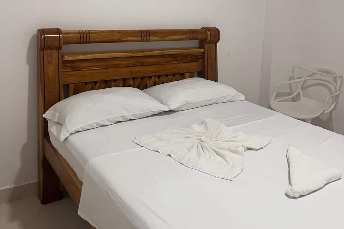 Una cama bien hecha con sábanas blancas y cabecera de madera en una habitación sencilla con paredes y una silla blancos.