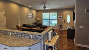 A modern living room with a kitchen area, a ceiling fan, and a large window with blinds. - Luxurious Amenities at Zoey's House in Heart of Olivette (Olivette)