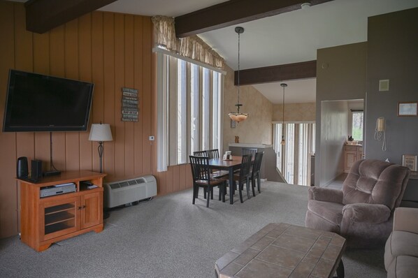 A living room with a television, wooden panel walls, a dining table with chairs, a sofa, and a large window. - Villa Como at Interlaken |Short Walk to Beach |Pool |Gas Fireplace |Ski at Alpine or Grand Geneva (Lake Geneva)