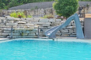 A swimming pool with a slide, surrounded by stone walls and a grassy area.