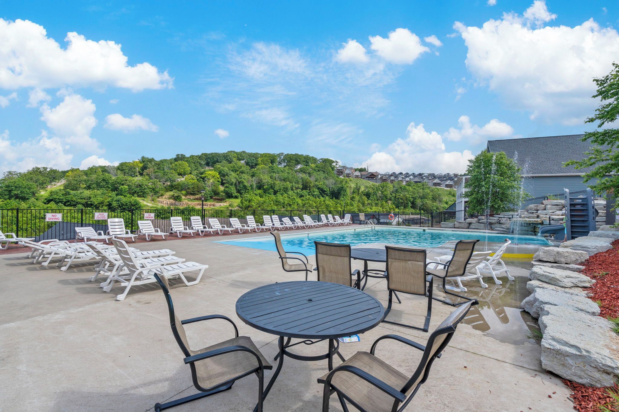 An outdoor pool area with lounge chairs, a table, and a view of a hill with trees and buildings.