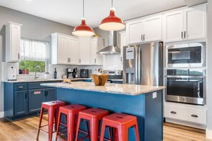 Une cuisine moderne avec des meubles blancs, un îlot central bleu, des tabourets de bar rouges, des appareils en acier inoxydable et des luminaires suspendus.